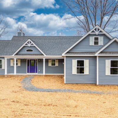 Custom-built Craftsman-style home with blue siding, white shutters, and a purple front door, surrounded by a gravel pathway and bare landscape, showcasing the craftsmanship of Craftsman Custom Homebuilders.